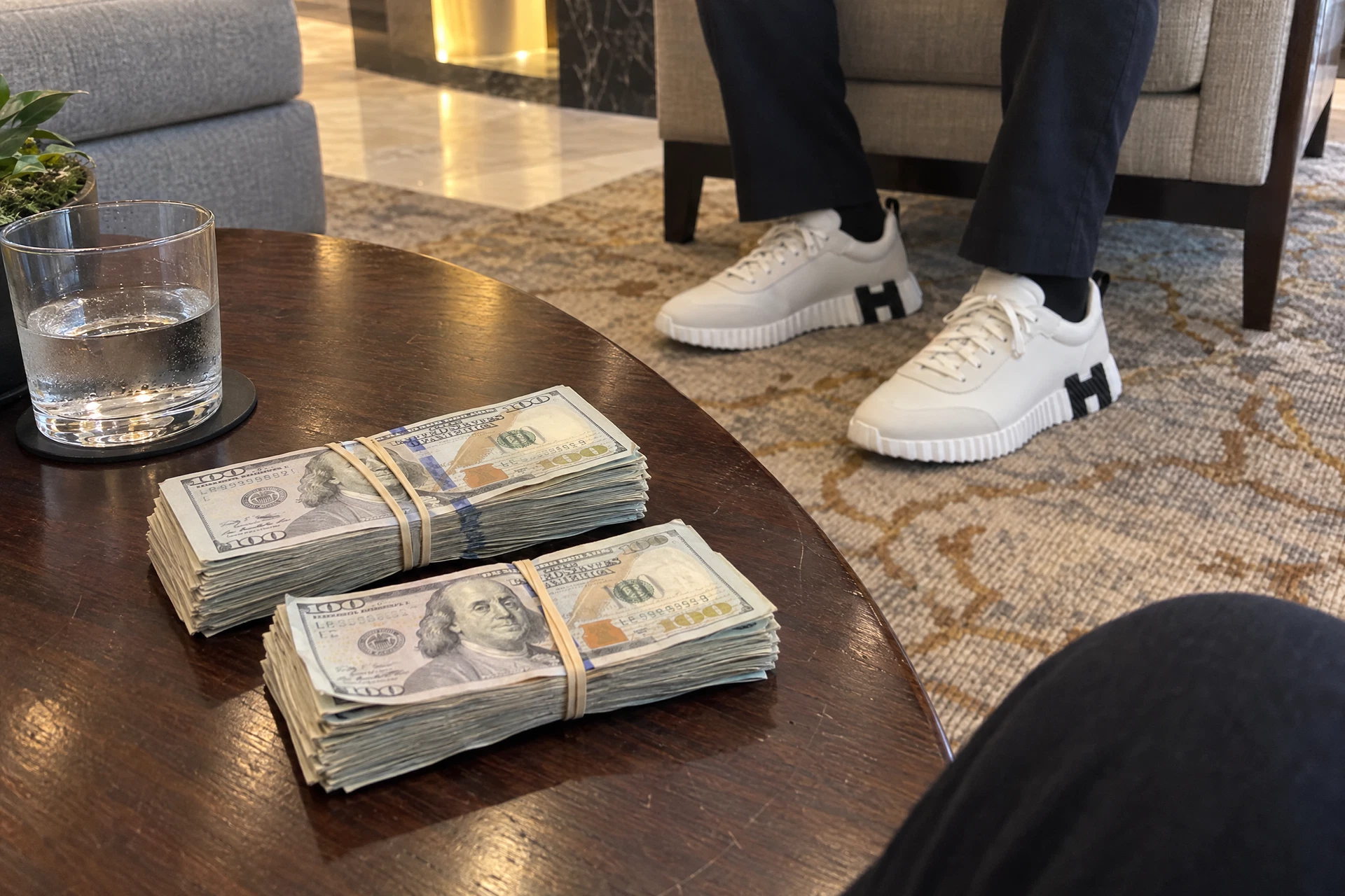 Illustrative scene of US dollars bundles held with rubber bands on a hotel-lobby table with a seated person’s sneakers visible